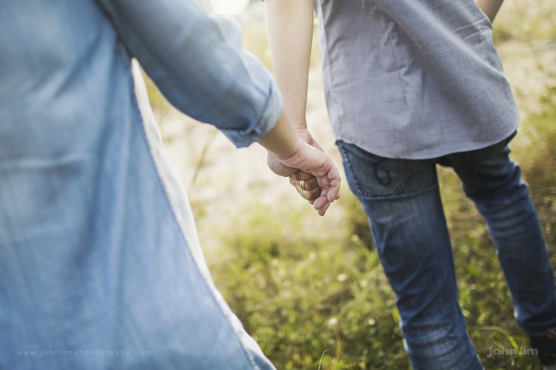 outdoor grass field maternity photos, parents of newborn baby holding hands with sun flare and wedding rings