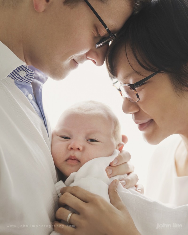 Newborn Studio Photography in Singapore, newborn baby boy held lovingly between parents, Daddy and Mummy looking lovingly at baby with natural window light